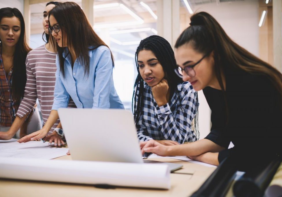 Four women collaborating on a project around a laptop and papers in an office setting.