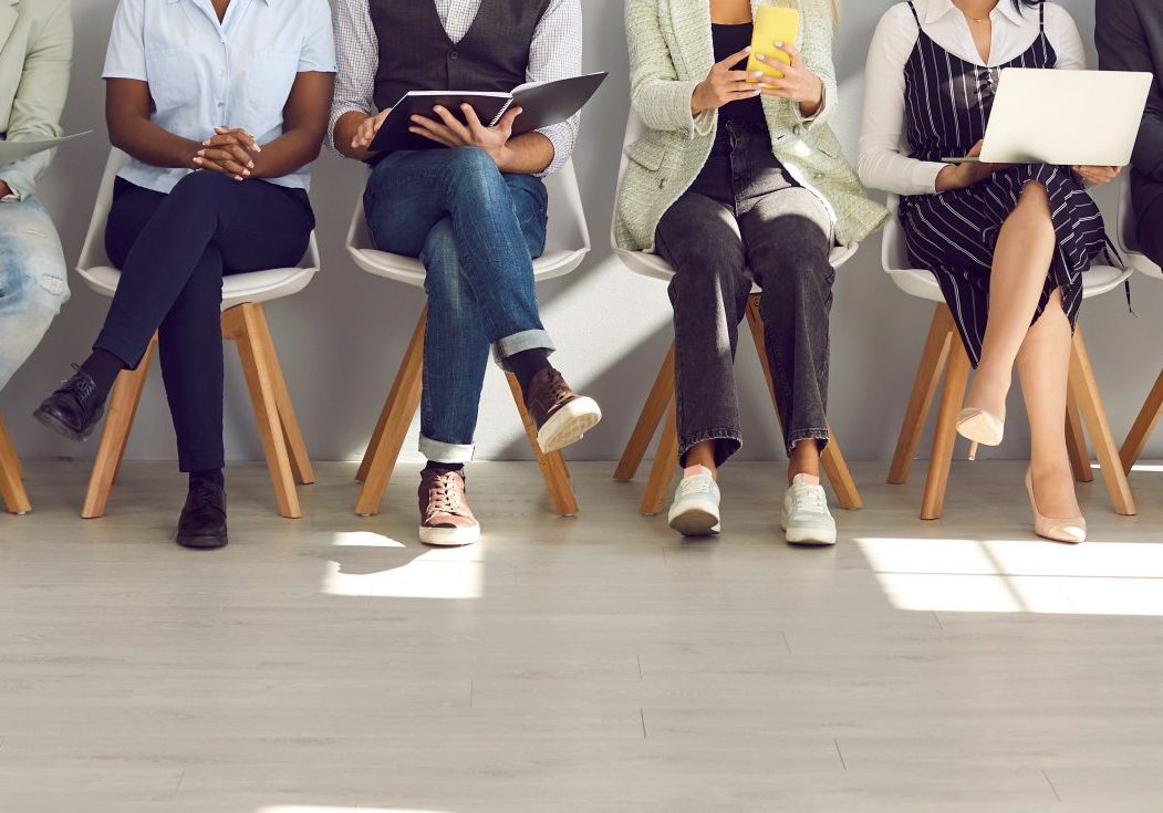 A row of people sitting on chairs, legs crossed, holding various electronic devices