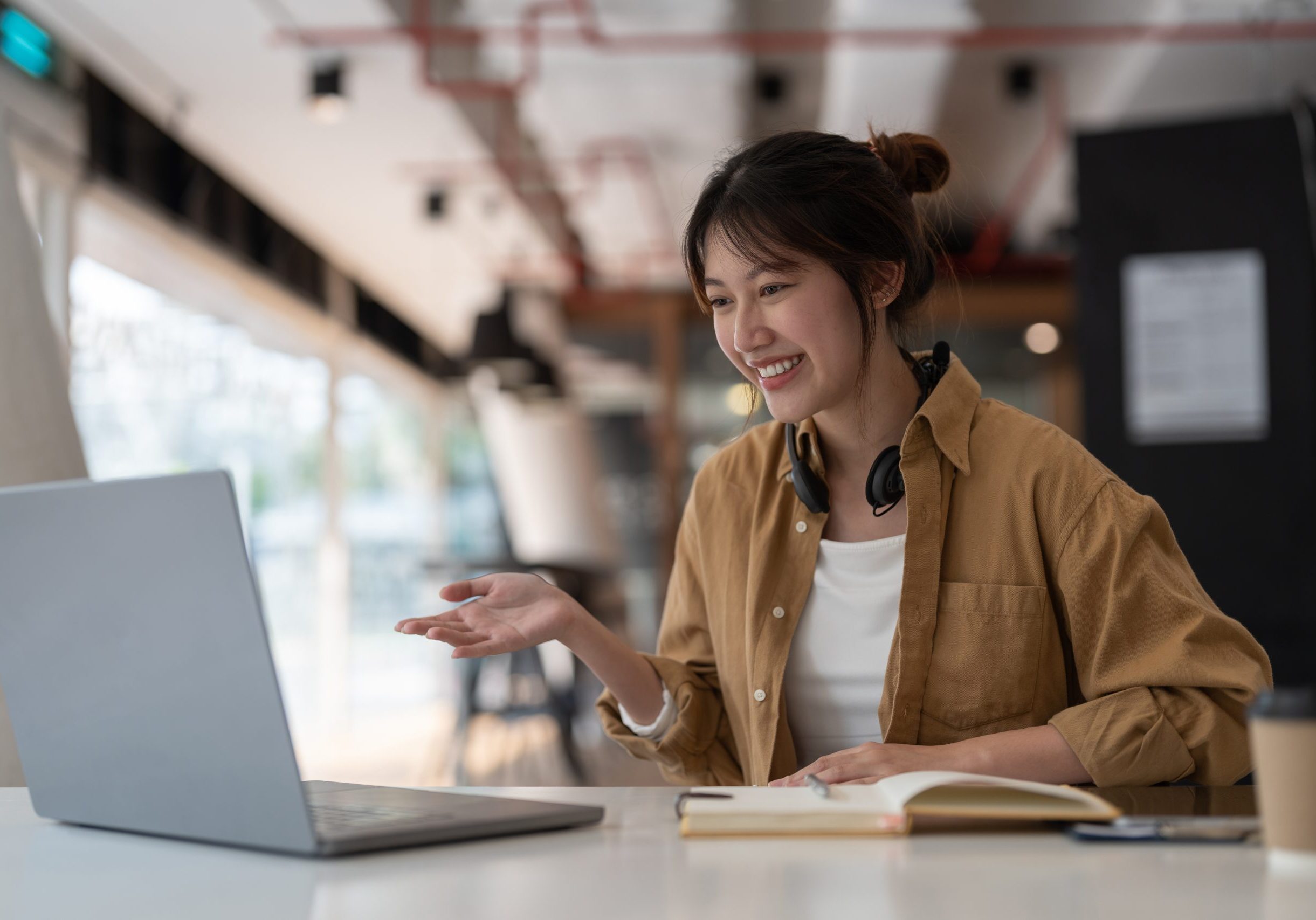 A young woman smiles while gesturing towards a laptop screen in a modern office setting, with headphones around her neck and a notebook beside her.